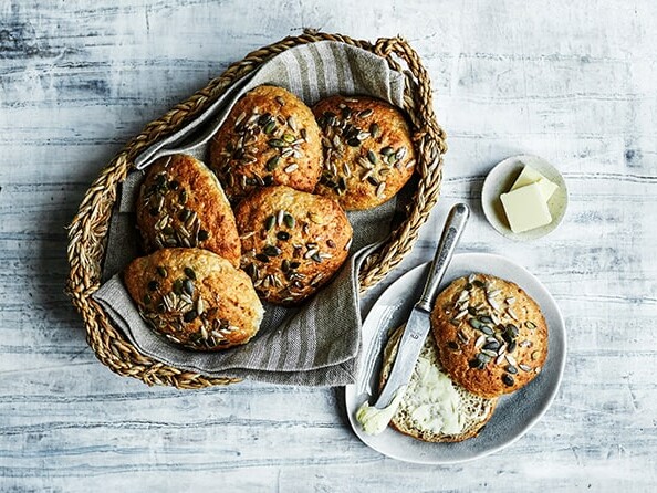 En kurv med boller toppet med frø og en tallerken med smør og en kniv.