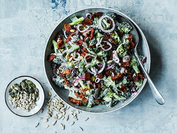 En skål fyldt med broccolisalat med bacon og rødløg, ved siden af frø.