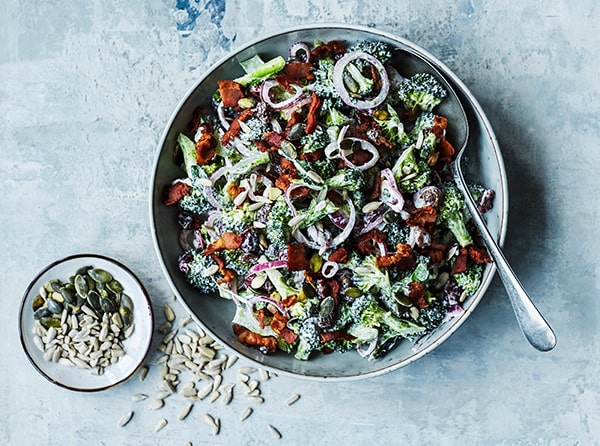 En skål fyldt med broccolisalat med bacon og rødløg, ved siden af frø.