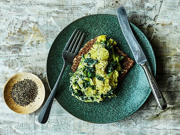 En grøn tallerken med rugbrød toppet med avokado og grøntsager ved siden af en gaffel og kniv.