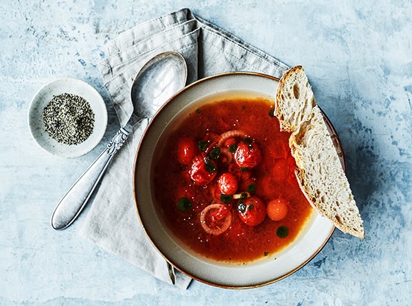 En skål tomatsuppe med cherrytomater, brødskive og krydderi ved siden af