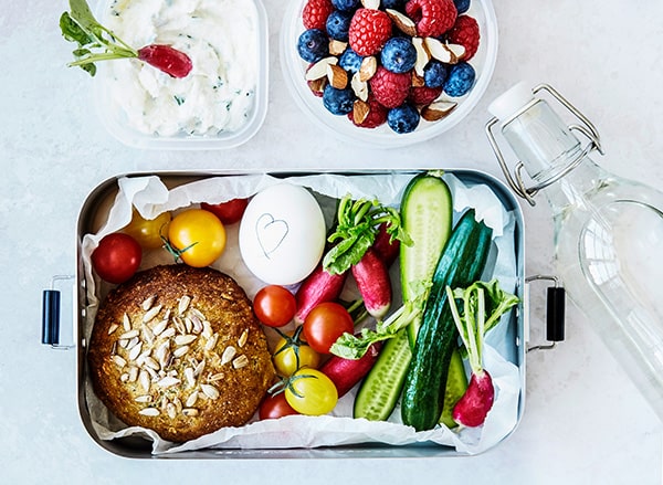 En frokostboks fyldt med grøntsager, æg, og en skål bær ved siden af