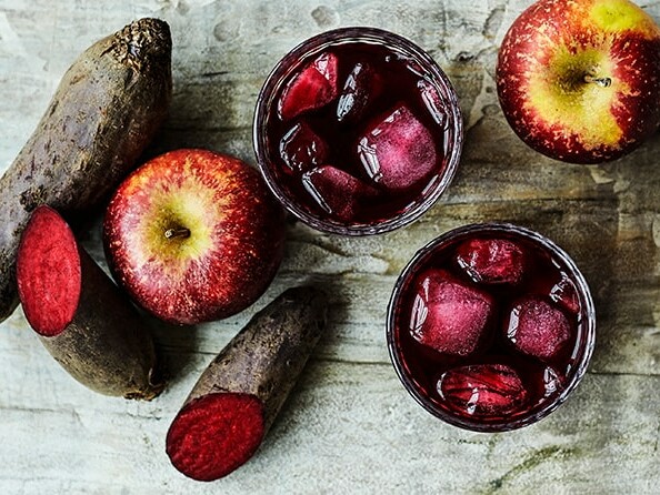 To glas rødbede- og æblejuice med isterninger, omgivet af frisk rødbede og æbler.