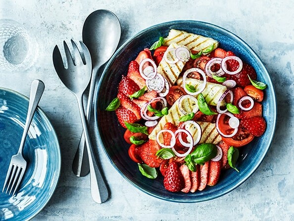 En farverig skål med frugt og grøntsager, inklusiv jordbær, tomater og grillet ost, på et blåt fad