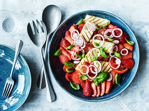 En farverig skål med frugt og grøntsager, inklusiv jordbær, tomater og grillet ost, på et blåt fad