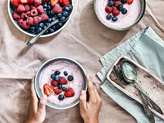 Skåle med yoghurt og friske bær på et bord med lyserødt stof.