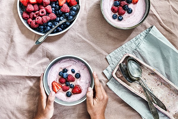 Skåle med yoghurt og friske bær på et bord med lyserødt stof.