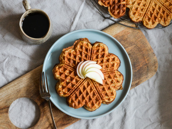 En tallerken med vafler toppet med æbleskiver ved siden af en kop kaffe.