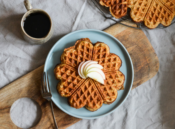 En tallerken med vafler toppet med æbleskiver ved siden af en kop kaffe.