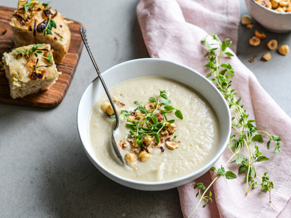 En skål rodfrugtsuppe pyntet med nødder og urter, ved siden af brød
