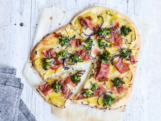 Hjerteformet pizza med kartofler, skinke, broccoli og løg på hvidt træbord.