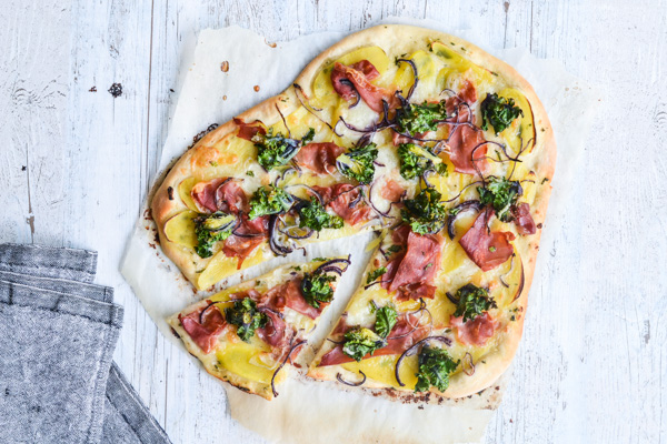 Hjerteformet pizza med kartofler, skinke, broccoli og løg på hvidt træbord.