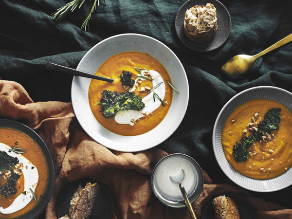 Tre skåle græskarsuppe med creme fraiche og grønt pynt, serveret med brød.