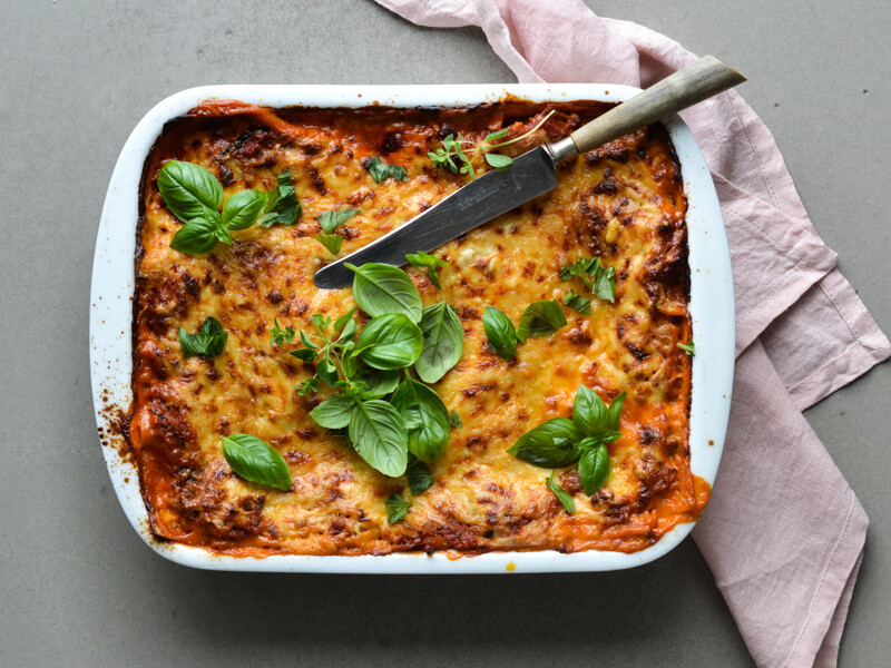 Lasagne i en hvid fad dekoreret med frisk basilikum og en kniv på toppen