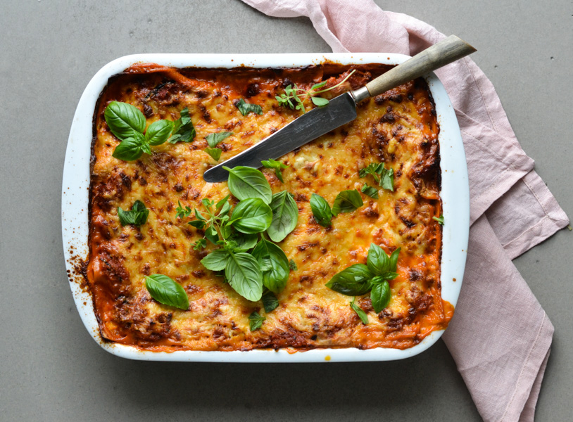 Lasagne i en hvid fad dekoreret med frisk basilikum og en kniv på toppen