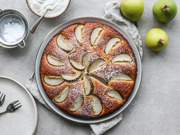 Friskbagt pærekage med flormelis, omgivet af pærer, flormelis og en tallerken