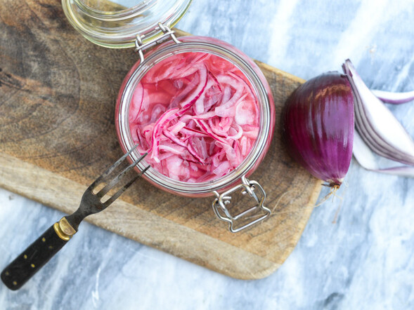 Et glas med syltede rødløg på et skærebræt ved siden af en hel rødløg
