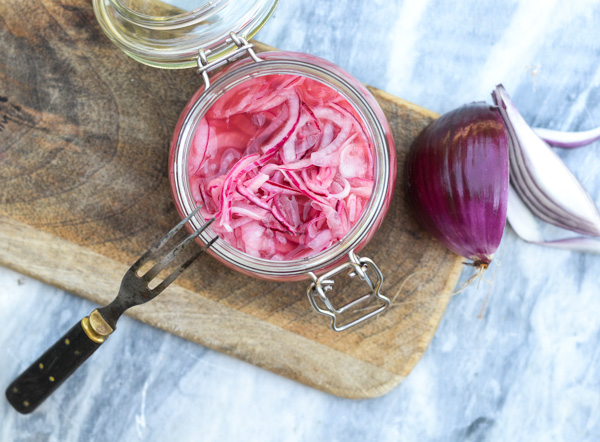 Et glas med syltede rødløg på et skærebræt ved siden af en hel rødløg