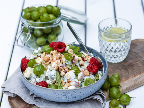 Skål med salat toppet med stikkelsbær, hindbær og nødder ved siden af et glas vand og krukke med stikkelsbær.