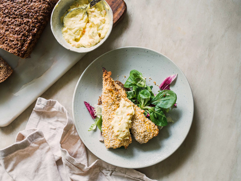 En tallerken med sprød fiskefilet, remoulade og grøn salat. Ved siden af ligger rugbrød samt en serviet.
