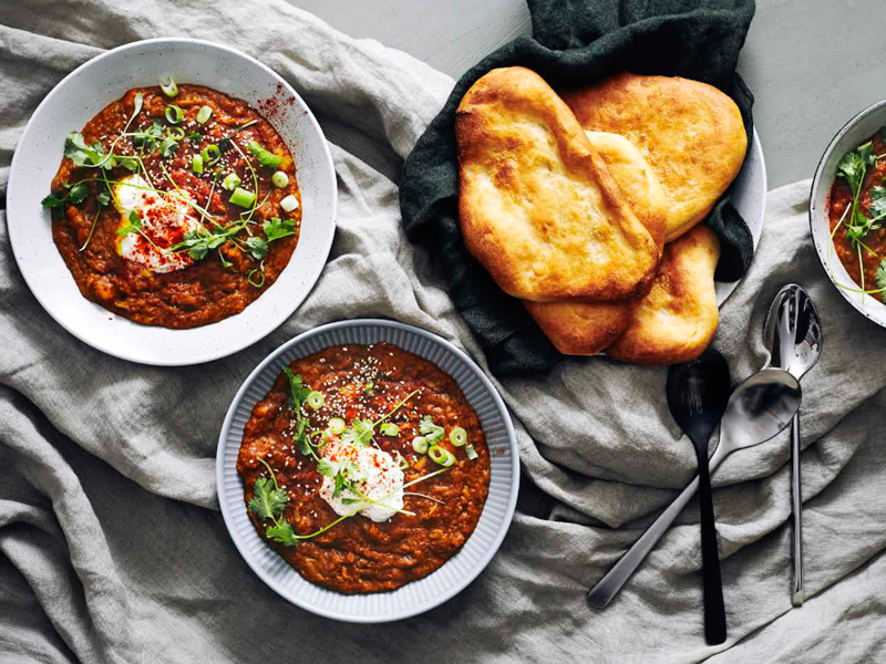 To skåle med krydret gryderet, pyntet med cremefraiche og urter, ved siden af fladbrød