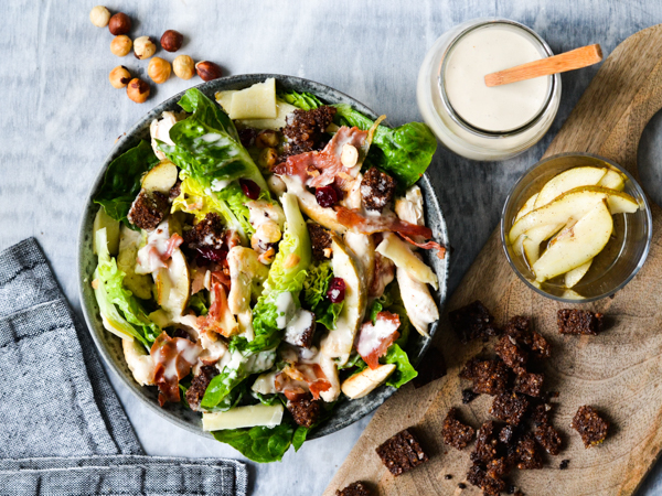 Salatskål med kylling, brødcroutoner og nødder, ledsaget af dressing og pæreskiver på skærebræt
