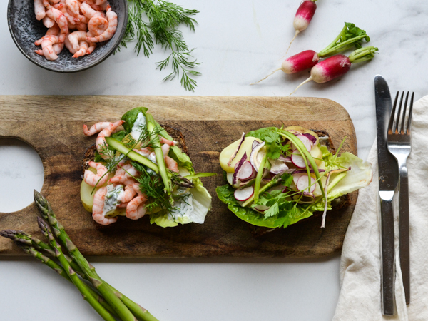 To smørrebrød på skærebræt med rejer, asparges og radiser.
