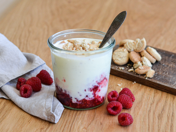 Glas med hindbær trifli og en ske, omringet af friske hindbær og knuste småkager.