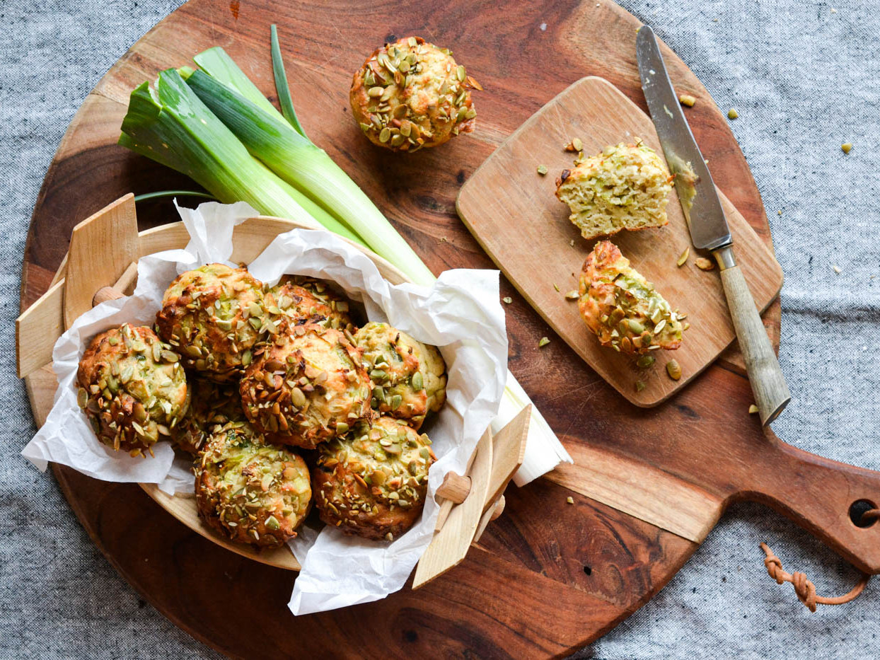 Hjemmelavede muffins med flækkede græskarkerner, porre ved siden af og en brødkniv på et skærebræt.