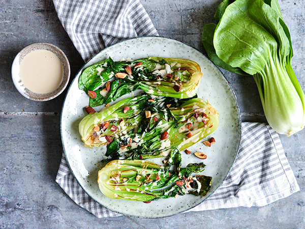 En tallerken med bagt pak choi toppet med mandler, med en skål dressing ved siden af.